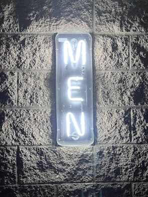 A neon sign glowing brightly against a textured stone wall, displaying the word 'MEN' in vertical alignment with white light. The illumination casts a soft glow around the sign, highlighting the rough surface of the wall.