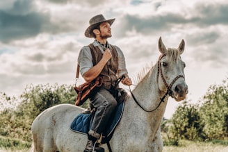 A man in a cowboy-style outfit sits astride a white horse in an open field. The rider wears a brown hat, vest, and blue jeans, holding the reins confidently. The sky is partly cloudy, and the surrounding landscape is lush with greenery.