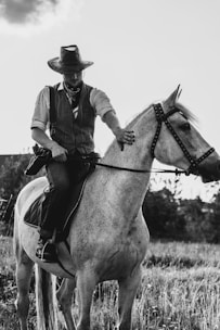 A person is riding a horse in a countryside setting, wearing a cowboy hat, a bandana, and a vest. The horse is light-colored and standing on a field with grass. The scene is captured in black and white, suggesting an old-fashioned or rustic atmosphere.