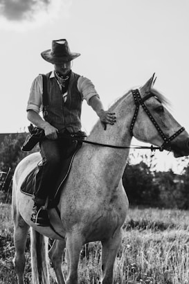 A person is riding a horse in a countryside setting, wearing a cowboy hat, a bandana, and a vest. The horse is light-colored and standing on a field with grass. The scene is captured in black and white, suggesting an old-fashioned or rustic atmosphere.