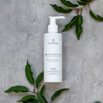 A white bottle of skincare product (BIO Gel Detergente Viso) with a pump dispenser is placed on a grey textured surface, surrounded by green leaves.
