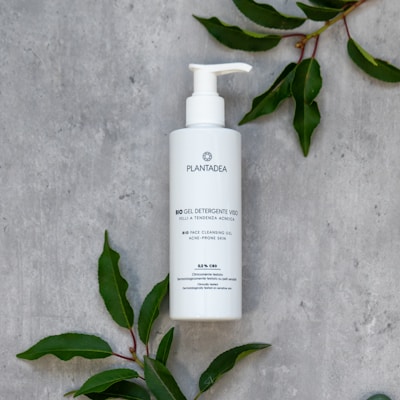 A white bottle of skincare product (BIO Gel Detergente Viso) with a pump dispenser is placed on a grey textured surface, surrounded by green leaves.