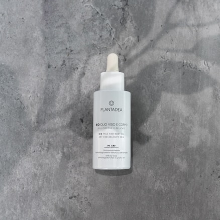 A white bottle with a dropper lid labeled as 'PlantaDea Bio Olio Viso e Corpo'. The bottle sits on a textured gray surface, casting a shadow.