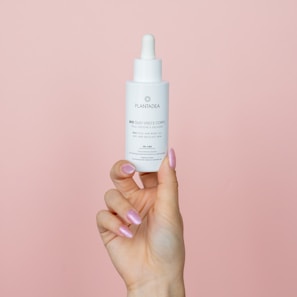 A hand with pink, glossy nails holds a white dropper bottle against a pale pink background. The bottle features text and a logo, related to skincare or cosmetic oil.