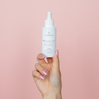 A hand with pink, glossy nails holds a white dropper bottle against a pale pink background. The bottle features text and a logo, related to skincare or cosmetic oil.