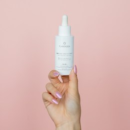 A hand with pink, glossy nails holds a white dropper bottle against a pale pink background. The bottle features text and a logo, related to skincare or cosmetic oil.