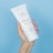 A hand holding a tube of Plantadea bio body cream against a solid light blue background. The hand has neatly manicured nails with a light pink nail polish. The tube is white with text indicating it is for dry and delicate skin, containing 1% CBD.