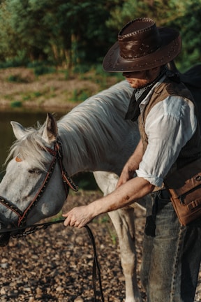 A person wearing a brown cowboy hat and vest is tending to a white horse standing on a rocky surface near a body of water. The setting appears natural, with green foliage in the background, and the person is holding the horse's reins.