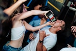 A lively scene at what appears to be a party, where a woman is pouring a drink into a man's mouth from a bottle. The man is gesturing with his hand in a playful manner, and there are several other people in the background. The atmosphere seems energetic and social.