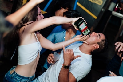 A lively scene at what appears to be a party, where a woman is pouring a drink into a man's mouth from a bottle. The man is gesturing with his hand in a playful manner, and there are several other people in the background. The atmosphere seems energetic and social.
