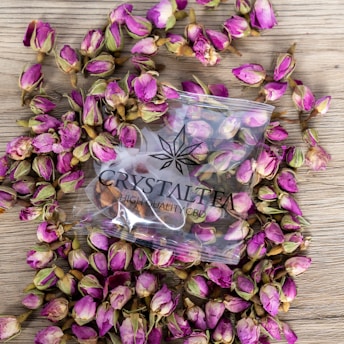 A collection of small pink rosebuds scattered on a wooden surface surrounds a clear plastic bag labeled 'CRYSTAL TEA HIGH QUALITY CBD'. The bag contains similar rosebuds and features a simple, modern logo design.