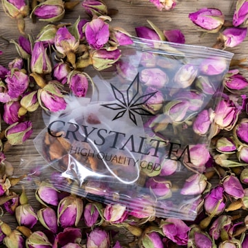A clear plastic bag labeled 'CRYSTAL TEA HIGH QUALITY CBD' is surrounded by numerous dried pink rosebuds on a wooden surface. The bag is transparent, allowing the contents and the decorative rosebuds beneath to be seen clearly.
