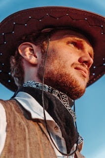 A person is depicted wearing a brown cowboy hat, a black bandana with white patterns around the neck, and a textured brown vest over a white shirt. The background features a clear blue sky suggesting an outdoor setting.