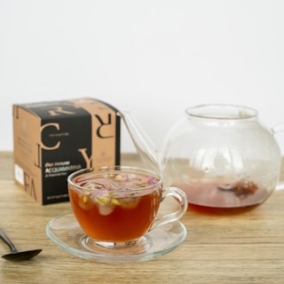 A clear glass teacup filled with a reddish beverage, garnished with small pink flowers, is placed on a wooden surface. Next to the cup is a matching glass teapot partially filled with the same beverage. A black spoon lies on the table, and there is a box labeled 'ACQUAMARINA' in the background.