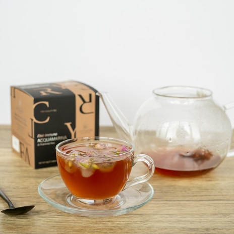 A clear glass teacup filled with a reddish beverage, garnished with small pink flowers, is placed on a wooden surface. Next to the cup is a matching glass teapot partially filled with the same beverage. A black spoon lies on the table, and there is a box labeled 'ACQUAMARINA' in the background.