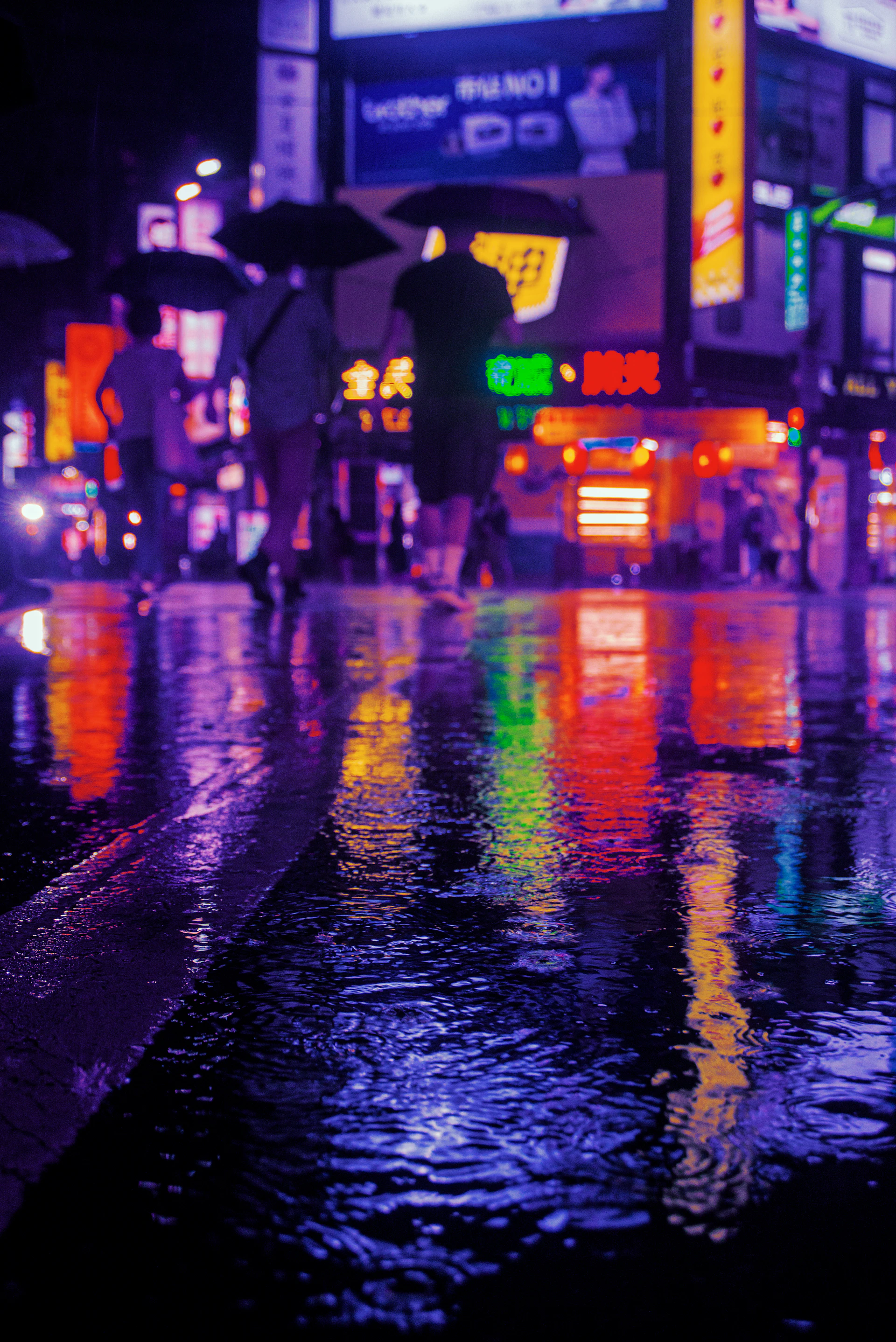 An urban street scene at night, with colorful neon signs reflecting on wet pavement and blurred motion of passing pedestrians.