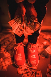 A person is holding a large amount of US dollars in their hands, wearing red shoes with white lettering, standing on a floor scattered with more dollar bills. The image has a warm, reddish-orange lighting.