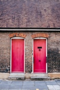 Two red doors set within a brick building facade, each with unique door hardware and steps leading up to them. The roof above is covered in dark brown shingles.