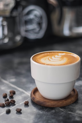 A cup of coffee with latte art on top sits on a wooden coaster. Coffee beans are scattered nearby on a marble surface, providing a rustic and cozy ambiance. In the blurred background, other coffee-making equipment is visible.