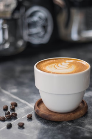 A cup of coffee with latte art on top sits on a wooden coaster. Coffee beans are scattered nearby on a marble surface, providing a rustic and cozy ambiance. In the blurred background, other coffee-making equipment is visible.