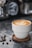 A cup of coffee with latte art on top sits on a wooden coaster. Coffee beans are scattered nearby on a marble surface, providing a rustic and cozy ambiance. In the blurred background, other coffee-making equipment is visible.