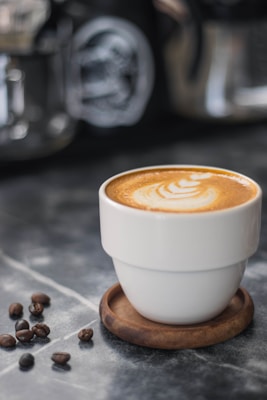 A cup of coffee with latte art on top sits on a wooden coaster. Coffee beans are scattered nearby on a marble surface, providing a rustic and cozy ambiance. In the blurred background, other coffee-making equipment is visible.