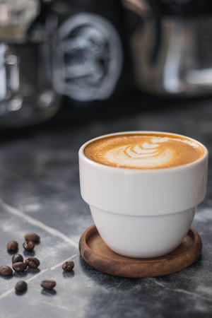 A cup of coffee with latte art on top sits on a wooden coaster. Coffee beans are scattered nearby on a marble surface, providing a rustic and cozy ambiance. In the blurred background, other coffee-making equipment is visible.