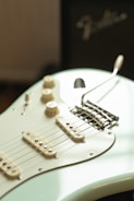 A close-up view of an electric guitar with a light cream body, showcasing the strings, pickups, and control knobs. The guitar is positioned against a blurred background with part of an amplifier visible, featuring a recognizable brand logo.