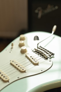 A close-up view of an electric guitar with a light cream body, showcasing the strings, pickups, and control knobs. The guitar is positioned against a blurred background with part of an amplifier visible, featuring a recognizable brand logo.