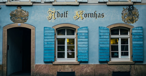 An ornate facade of a building features two windows with blue shutters and an arched doorway. The facade is adorned with intricate decorative elements and the name 'Adolf Kornhas' in stylized gold lettering. Coats of arms are painted on either side of the name.