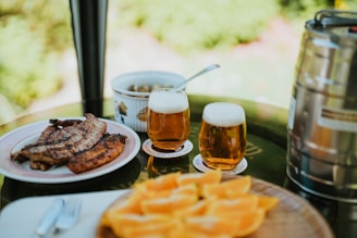 A table is set outdoors with plates of grilled meat and a bowl of olives. Two glasses of beer sit on coasters next to a metal keg. In the foreground, there is a platter of sliced fruit, possibly oranges.