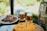 A table is set outdoors with plates of grilled meat and a bowl of olives. Two glasses of beer sit on coasters next to a metal keg. In the foreground, there is a platter of sliced fruit, possibly oranges.