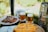 A table is set outdoors with plates of grilled meat and a bowl of olives. Two glasses of beer sit on coasters next to a metal keg. In the foreground, there is a platter of sliced fruit, possibly oranges.