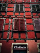 A close-up view of a building facade featuring decorative timber framing. The wall is painted in a vibrant red color with black wooden beams creating a grid-like pattern. There are multiple windows with closed wooden shutters painted in a darker shade of red. At the bottom of the image, there is a sign with text that reads 'L'Alchimiste' above a door or window.