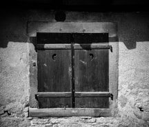A closed wooden window shutter with a textured stone wall surrounding it. The shutters feature a simple crescent-shaped cutout near the top.