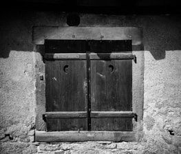 A closed wooden window shutter with a textured stone wall surrounding it. The shutters feature a simple crescent-shaped cutout near the top.