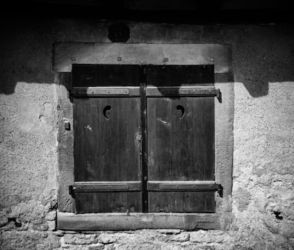 A closed wooden window shutter with a textured stone wall surrounding it. The shutters feature a simple crescent-shaped cutout near the top.