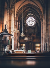 An ornate church interior featuring tall arches and a large circular stained glass window above an organ. The space includes chandeliers, wooden pews, and an altar adorned with flowers and religious symbols. Several people are seated and appear to be observing or participating in a church service.