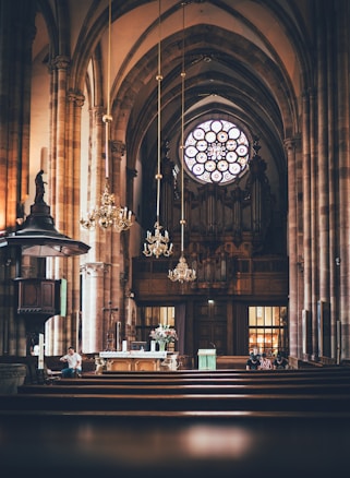 An ornate church interior featuring tall arches and a large circular stained glass window above an organ. The space includes chandeliers, wooden pews, and an altar adorned with flowers and religious symbols. Several people are seated and appear to be observing or participating in a church service.