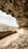 A rocky cave with textured walls opens up to an outdoor view of hills and shrubbery. Sunlight illuminates the cave interior, highlighting the rough textures and shapes of the rock formation. In the foreground, a low barrier or wall runs along the edge, providing a boundary.