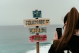 A colorful wooden sign with messages in Portuguese stands against the backdrop of the ocean, while a person with long hair takes a photo with a smartphone.