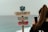 A colorful wooden sign with messages in Portuguese stands against the backdrop of the ocean, while a person with long hair takes a photo with a smartphone.
