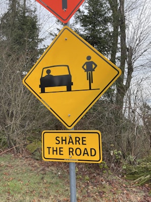 A yellow road sign is shown featuring a silhouette of a bicycle and a car, indicating the need for drivers and cyclists to share the road. Below it, there is an additional rectangular yellow sign with the text 'SHARE THE ROAD' in black letters. The signs are set against a backdrop of trees and foliage.