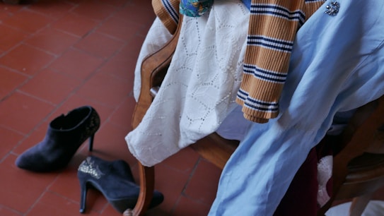 A wooden chair is draped with various clothing items, including a ribbed, striped sweater, a blue dress, and a white lace garment. On the floor nearby, a pair of black high-heeled boots with decorative details can be seen. The floor is composed of reddish-brown tiles.