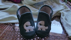 A pair of small blue baby shoes with white squirrel appliques displayed on a woven surface. In the background, a light-colored baby onesie with a playful design is partially visible.