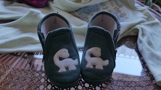 A pair of small blue baby shoes with white squirrel appliques displayed on a woven surface. In the background, a light-colored baby onesie with a playful design is partially visible.