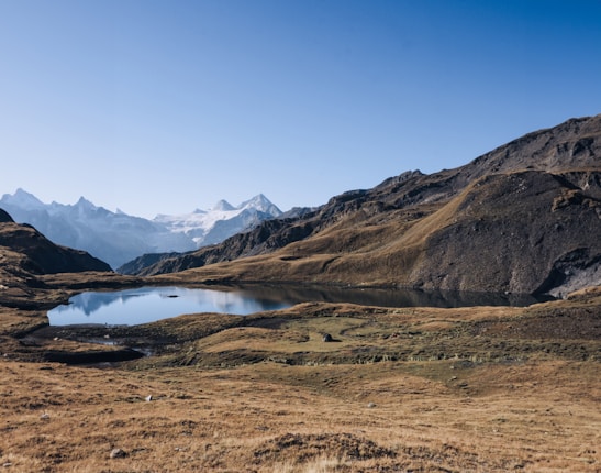 A tranquil mountain landscape featuring a small lake surrounded by rolling hills and rocky terrain. Snow-capped peaks rise in the distance under a clear blue sky, reflecting a serene and peaceful ambiance.