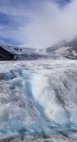 A vast, icy glacier stretches across the landscape under a partially cloudy sky. The ice is textured with ridges and bands of blue, indicating its density and age. Surrounding the glacier are dark rocky mountains, partially covered in snow.