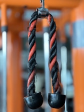 A close-up view of a twisted rope attachment with black and orange strands, commonly used in gym equipment for strength training exercises. The rope ends in black, rubberized grips, and it is suspended on a metallic hook, set against a blurred gym backdrop.