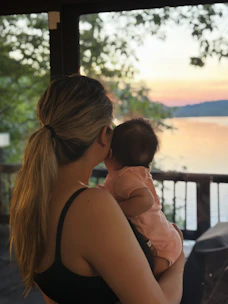 A serene mother holding her baby near a calm ocean shore at sunset.